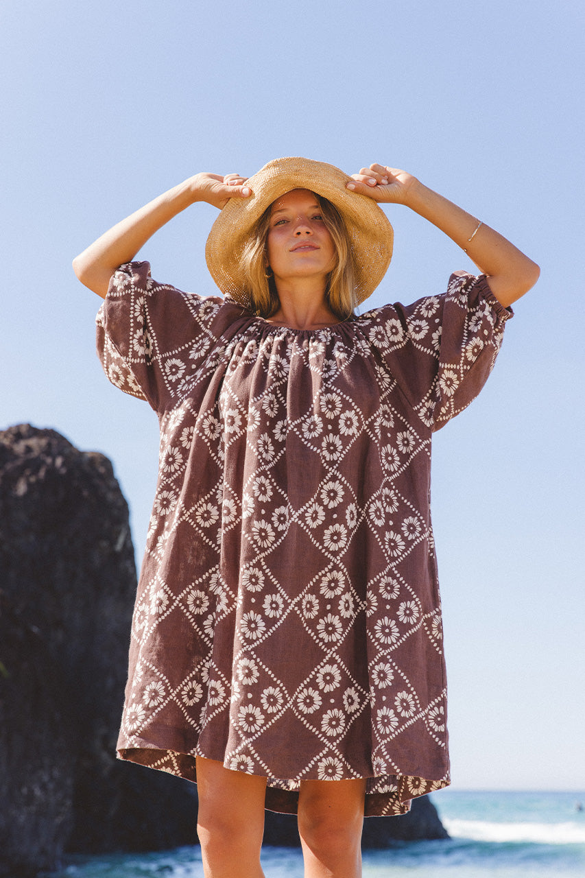 Dahlia Mini Dress Maple — beach with rocky headland, wide-brim hat styling, hand block print linen mini, maple brown geometric daisy pattern | Daughters of India