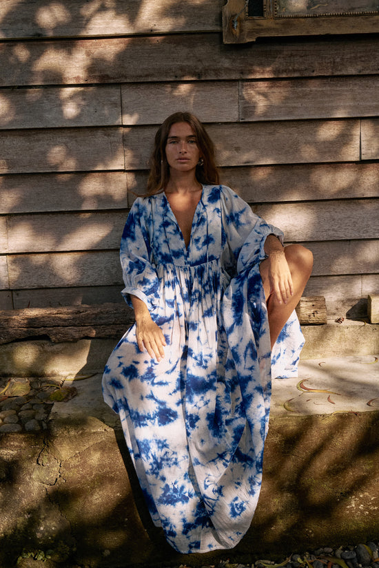 Kyra Maxi Dress Deep Sea — full length seated front view against rustic wooden wall, deep sea shibori tie-dye cotton maxi dress, relaxed flowing silhouette, dappled sunlight | Daughters of India