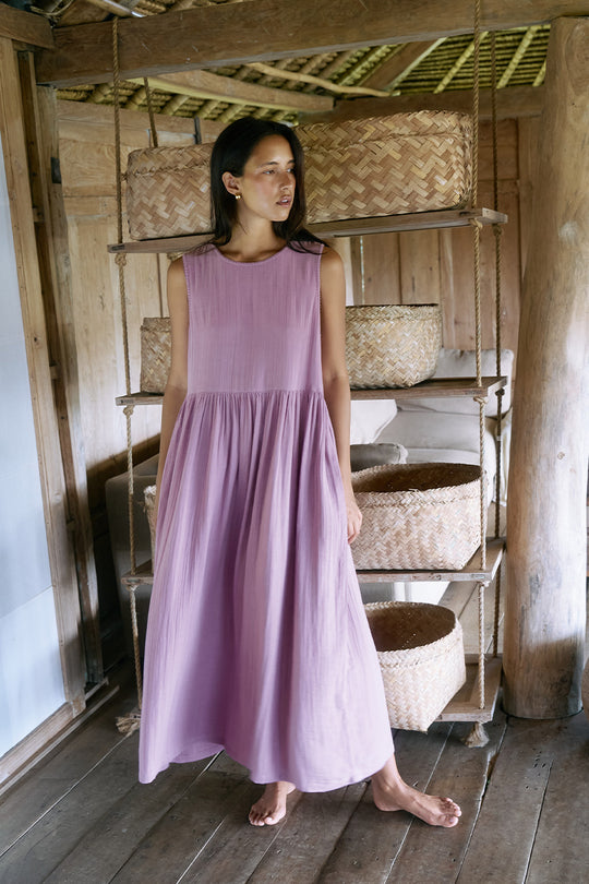 Nadi Dress Mauve — full length front view in rustic wooden interior with woven baskets, mauve double gauze cotton midi dress with gathered waist, natural bamboo shelving | Daughters of India